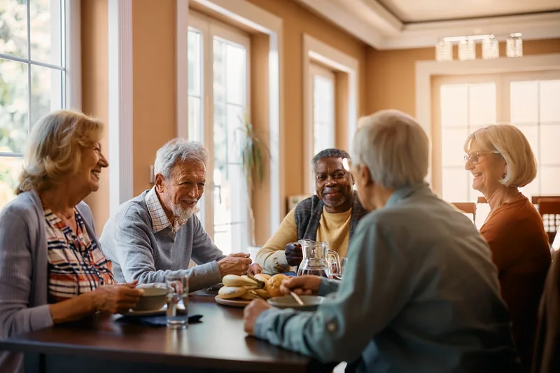 Residents talk and laugh over lunch at a table in a sunny dining room
