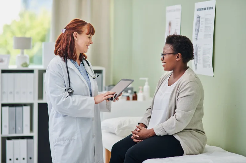 Clinician with a tablet talking with a woman seated on an exam table in a bright exam room