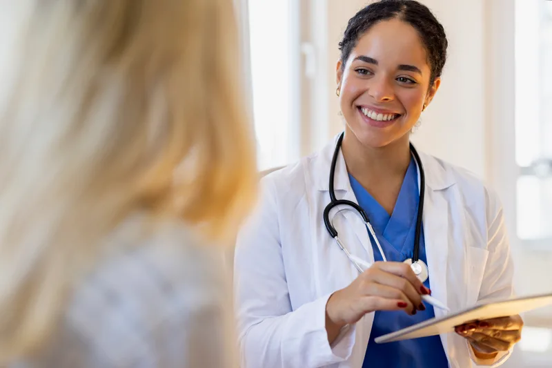 Smiling clinician in a white coat taking notes on a tablet while she listens to a patient
