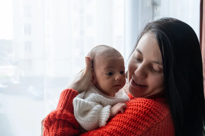 Mother in a red sweater holding her newborn close to her cheek by a sunny window