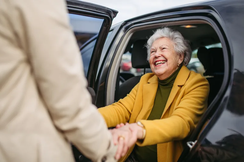 A smiling older woman in a mustard coat holds a helper's hands as she steps out of a car