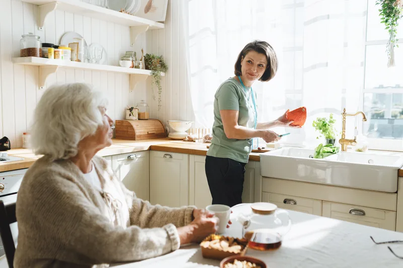 A caregiver cooks at the counter while an older woman sits at the kitchen table chatting with her