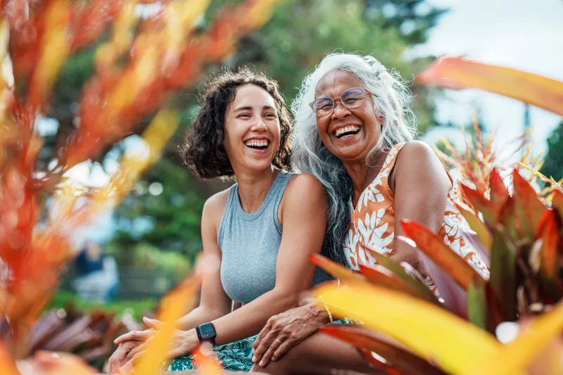 An adult daughter and her mother laugh together outdoors among bright plants