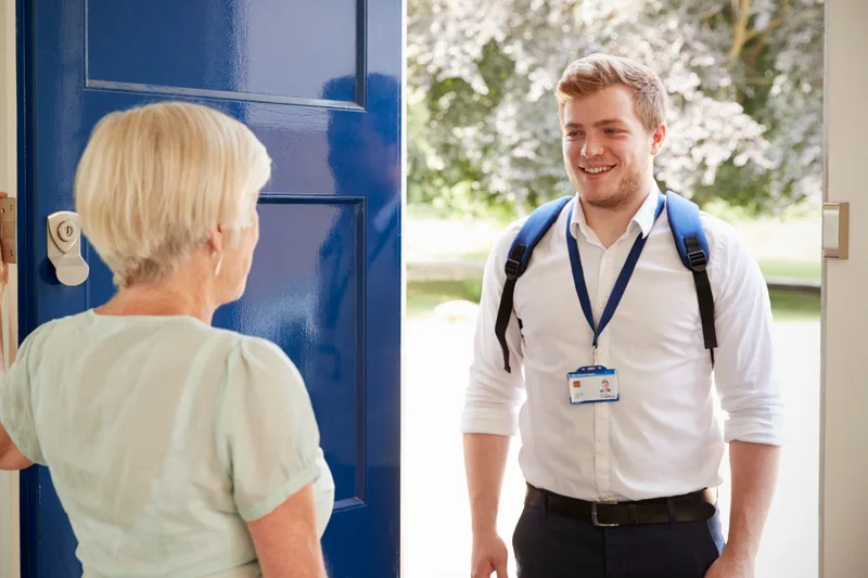 An older woman opens her front door to a smiling care worker with a lanyard and backpack
