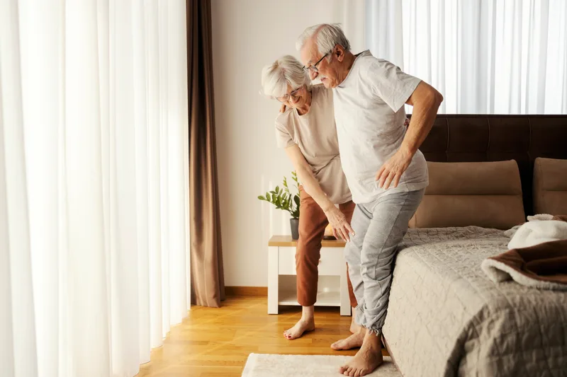 An older woman helps her husband stand up from the edge of the bed in their sunny bedroom