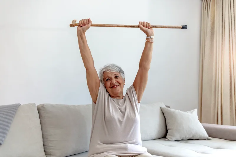 An older woman sitting on her couch raises her cane over her head with a big smile