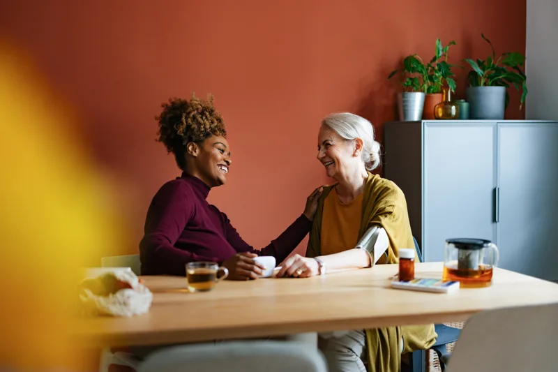 A caregiver and an older woman laugh together over cups of tea at a kitchen table