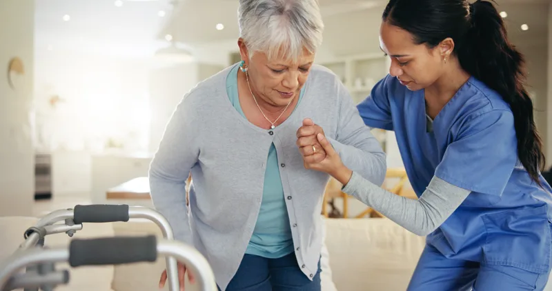 A caregiver steadies an older woman by the arm as she moves from her walker toward the couch