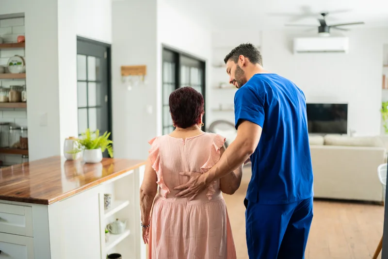 A male caregiver walks slowly beside an older woman through her bright living room, one hand at her back