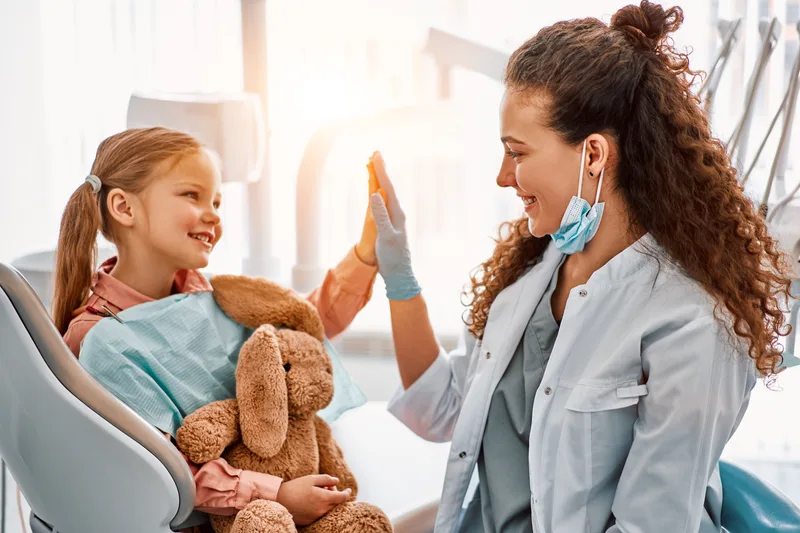 A pediatric dentist high-fives a smiling girl holding a stuffed bunny in the dental chair