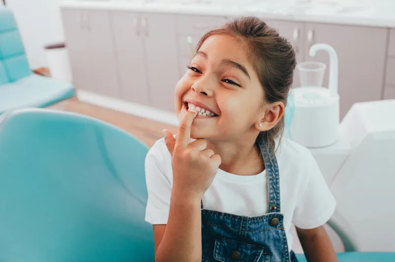 A smiling girl in a teal dental chair pointing at her teeth