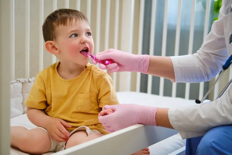 A toddler in a yellow shirt sitting on the exam chair while a dentist brushes his teeth