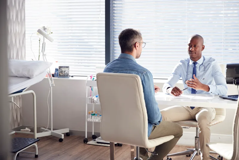 A physician talks with a patient at a sunlit exam-room desk, notes open between them