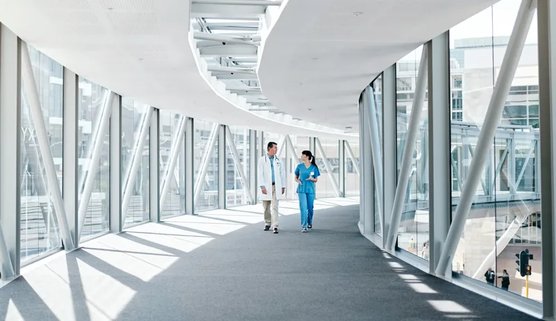 Two clinicians walk and talk in the glass corridor of a modern hospital