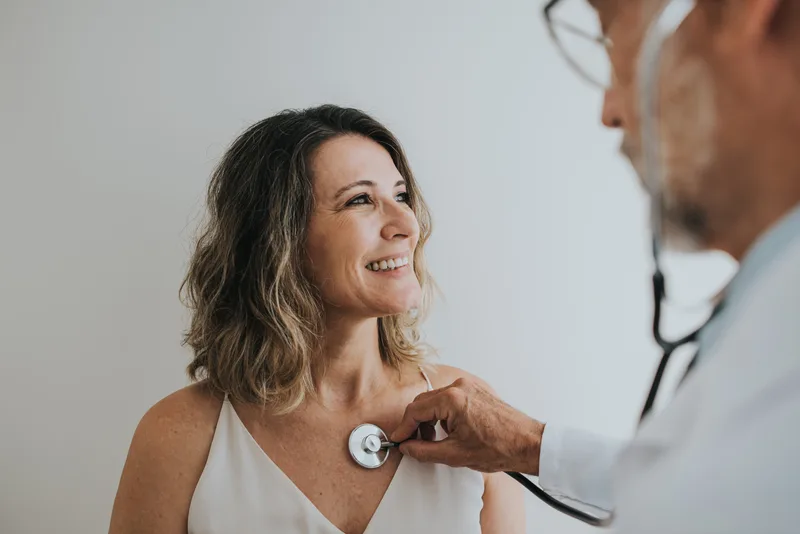 A smiling patient looks up at her cardiologist as he listens to her heart with a stethoscope