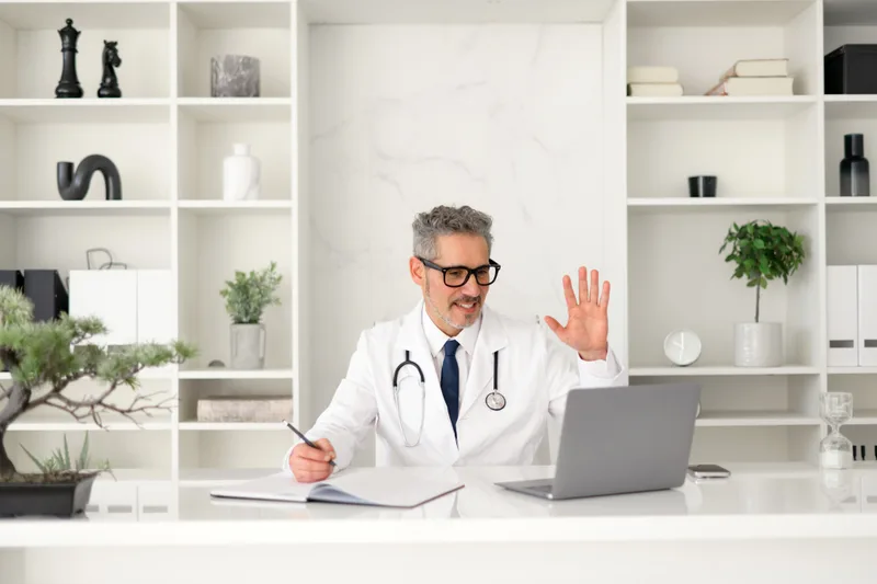 A grey-haired physician waves to a patient over a laptop video call from his desk