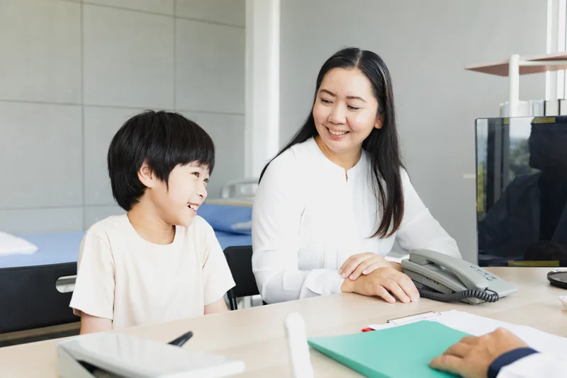 Smiling school-age boy at a clinic desk with his mother during a pediatric check-in