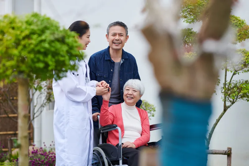 Riverbank clinician holding hands with a smiling older patient in a wheelchair in the hospital garden, her family beside her