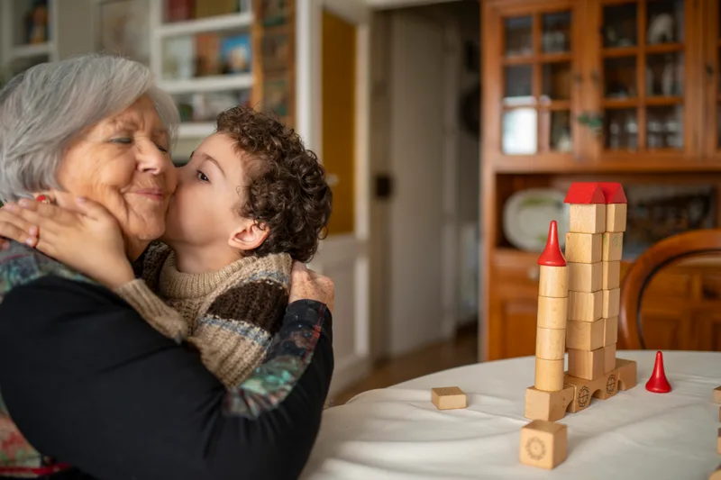 A grandmother laughs as her grandchild kisses her cheek at the family dining table