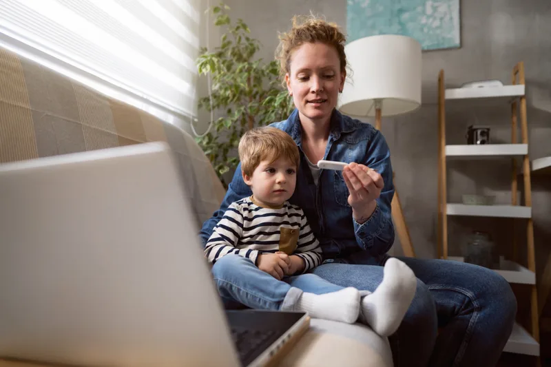 Mother on the sofa with her toddler on her lap, checking a thermometer during a video visit with the clinic