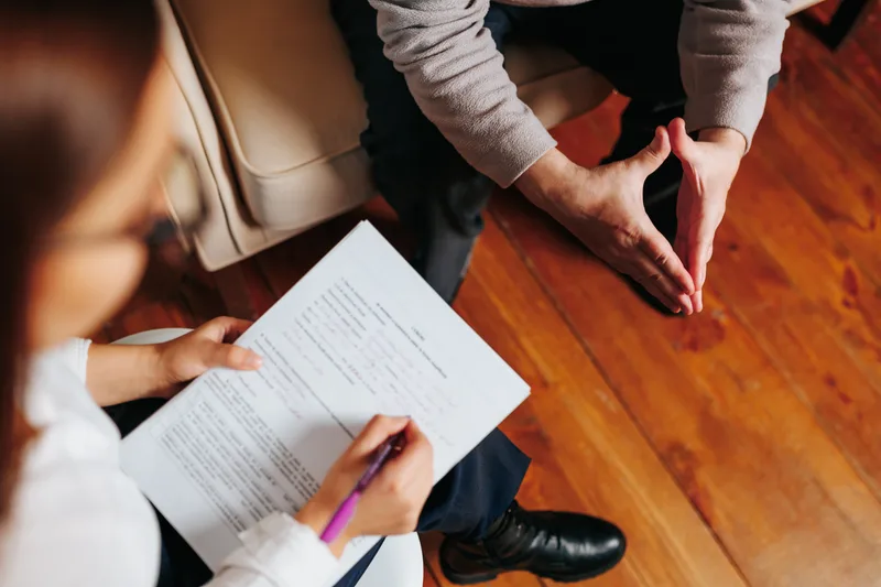 A clinician takes notes on a printed questionnaire while a patient sits across from her