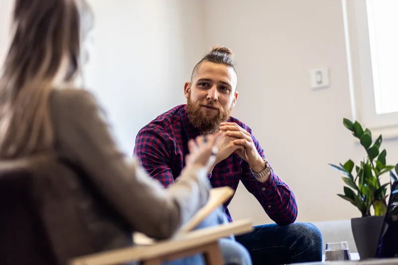A man in a plaid shirt leans forward and talks with a clinician in a bright office