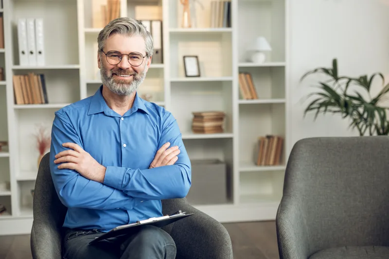 Dr. Graham Whitfield, a psychiatrist with grey hair and glasses, arms crossed in his office