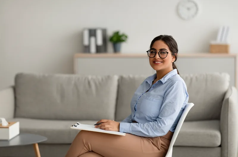 Leila Haddad, a psychiatric nurse practitioner with glasses, smiling while holding a clipboard
