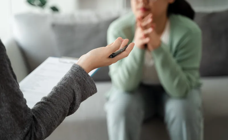 A clinician with a clipboard talks through options with a patient seated on a grey sofa