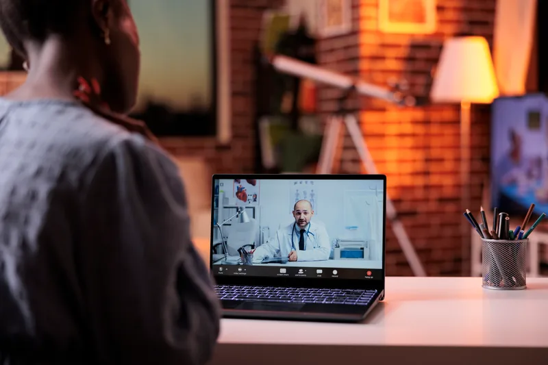 A patient joins an evening video visit from a desk at home, with the clinician on the laptop screen