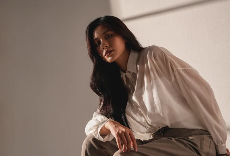 A woman in a soft linen shirt sits at home in warm window light, looking calm and relaxed