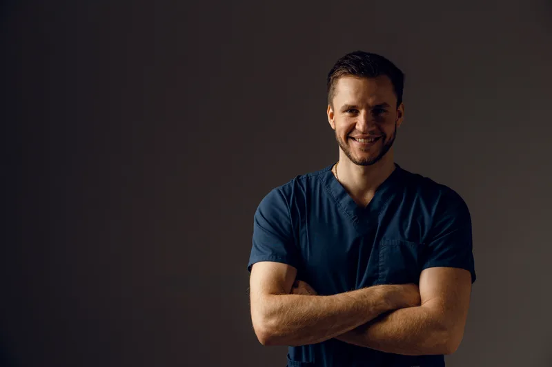 Portrait of a surgeon in navy scrubs with his arms crossed, smiling, against a dark wall