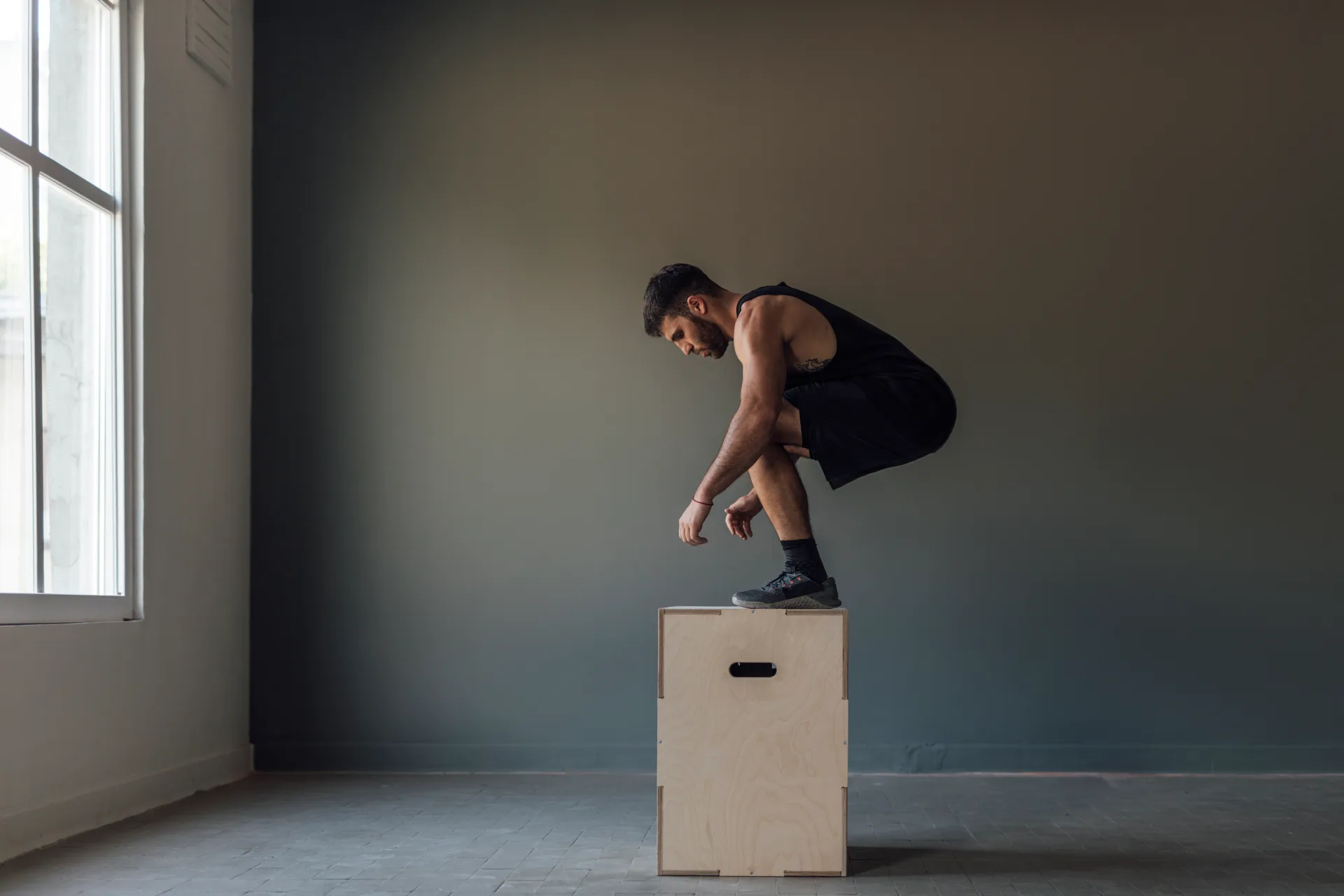 Athlete landing a jump on a plywood box in a minimalist grey training room lit by a window