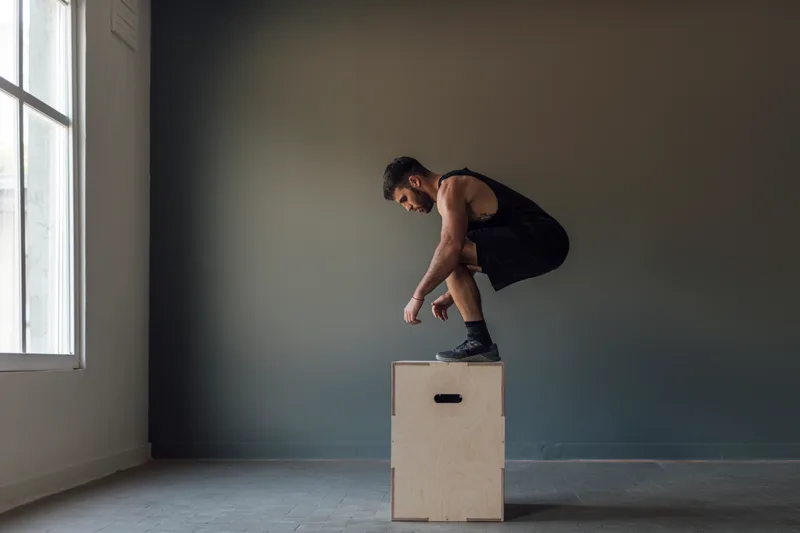 Athlete landing a jump on a plywood box in a minimalist grey training room lit by a window