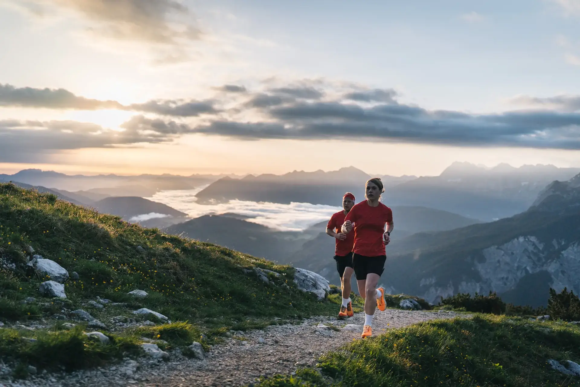 Two trail runners in red shirts climbing a mountain path above a sea of morning clouds