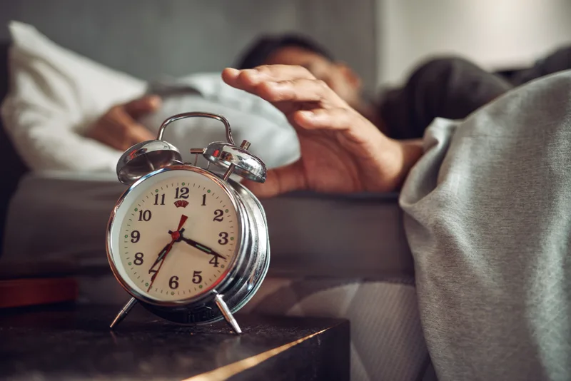 A man in bed reaching out to switch off a vintage alarm clock on his nightstand in early morning light