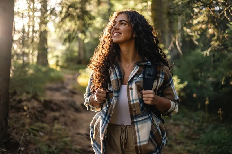 A smiling woman with curly hair hiking a sunlit forest trail, looking up at the trees