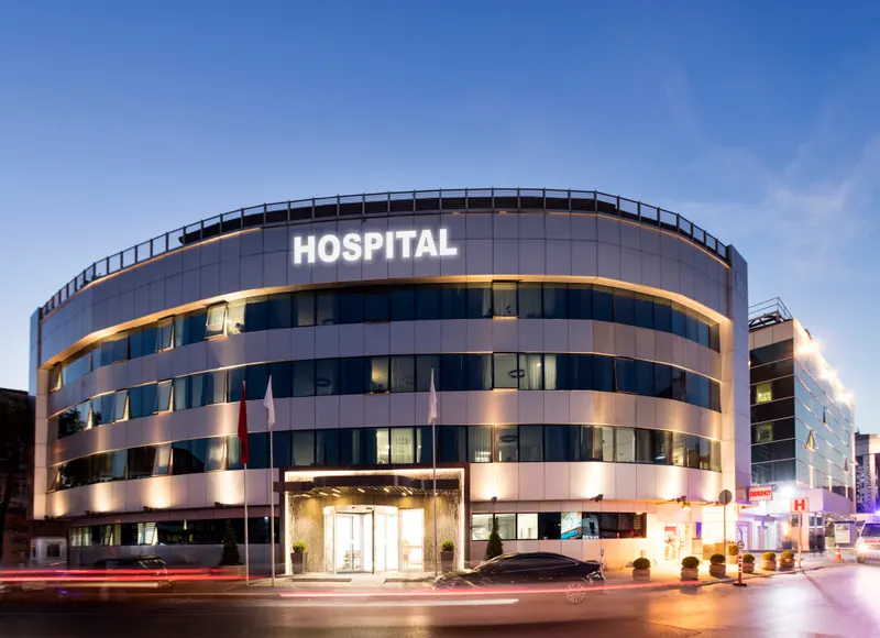 Curved glass-and-stone facade of Riverbank Medical Center lit at dusk, with a lighted hospital sign above the entrance canopy