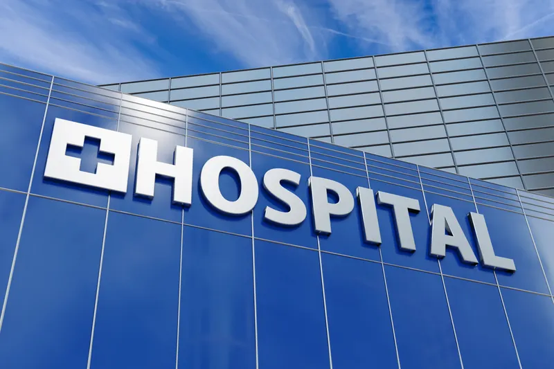 Blue glass curtain wall of Riverbank North Hospital with a white cross emblem and hospital lettering against a bright sky