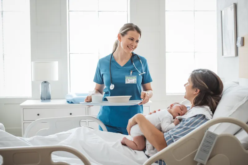 Nurse in teal scrubs bringing a meal tray to a mother cradling her sleeping newborn in a sunlit Family Birth Center suite