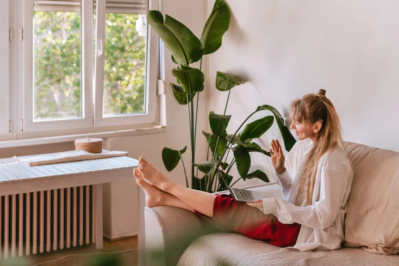 A woman with her feet up on the couch, waving hello to her therapist on a laptop video call