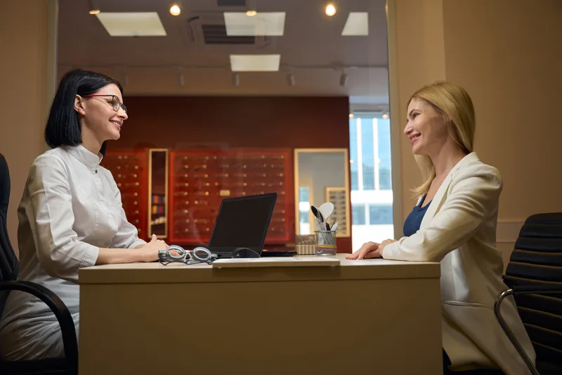 An optician talking a client through her options at a desk in front of the warm-lit frame wall
