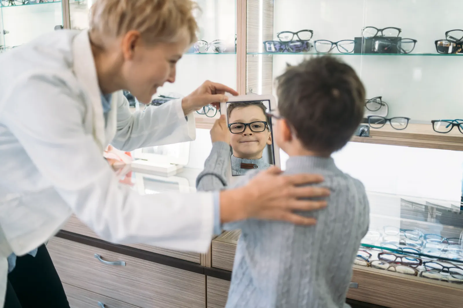 A boy checking his new glasses in a hand mirror while an optician steadies his shoulder