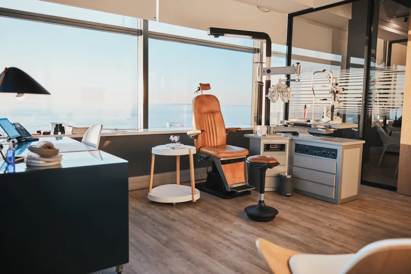 A bright exam room with a tan leather exam chair, a phoropter on its arm, and a slit lamp by the window