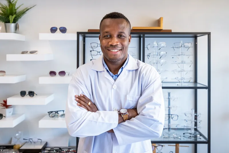 Marcus Osei, licensed optician, standing with his arms folded in front of the sunglasses shelves