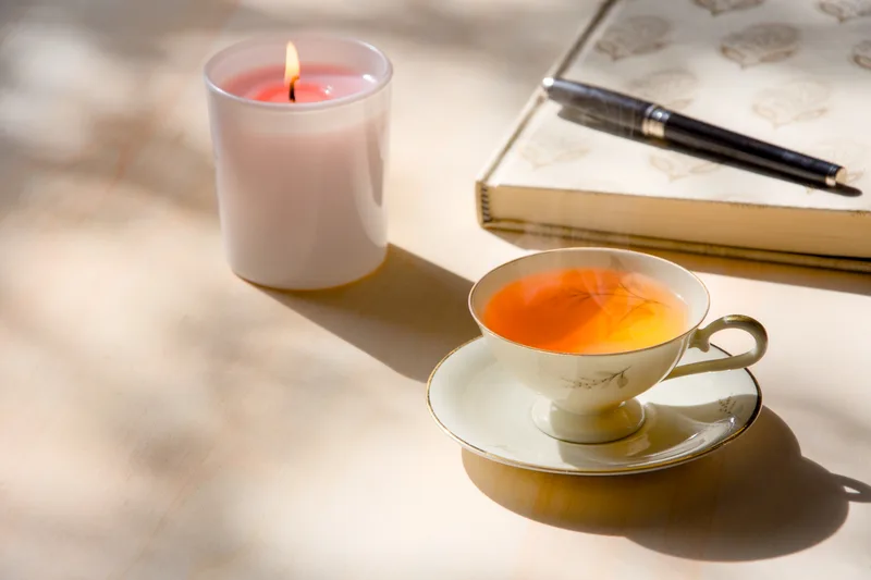 A cup of tea, a candle and a closed journal in soft afternoon light