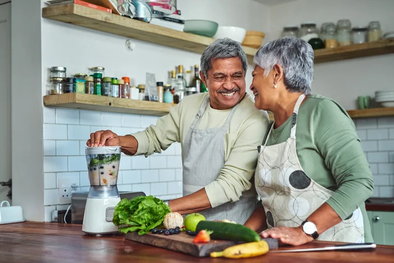 An older couple laugh while making a green smoothie in their kitchen