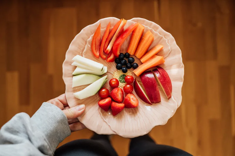 A hand holds a plate of sliced peppers, carrots, apples, strawberries and blueberries