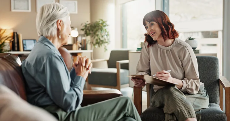 A clinician with a clipboard listens to an older woman in a calm, living-room style consult room