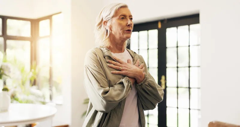 A woman in her fifties stands by a sunny window with her hands on her chest, taking a slow breath